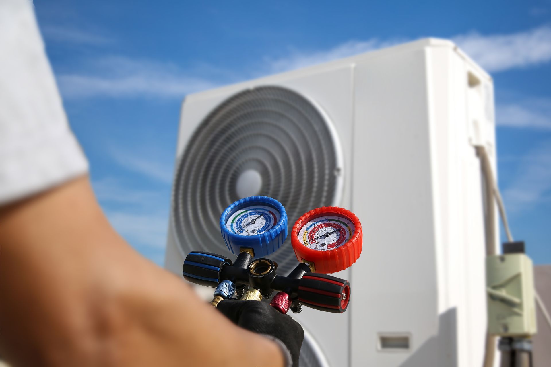 A technician holds a manifold gauge set in front of an outdoor air conditioning unit against a blue sky.