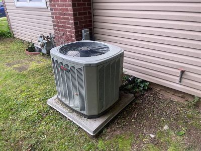 An air conditioning unit on a concrete pad near a brick wall and tan siding.