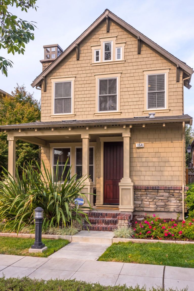 Tan two-story house with front porch, brick steps, and landscaping on a sunny day.