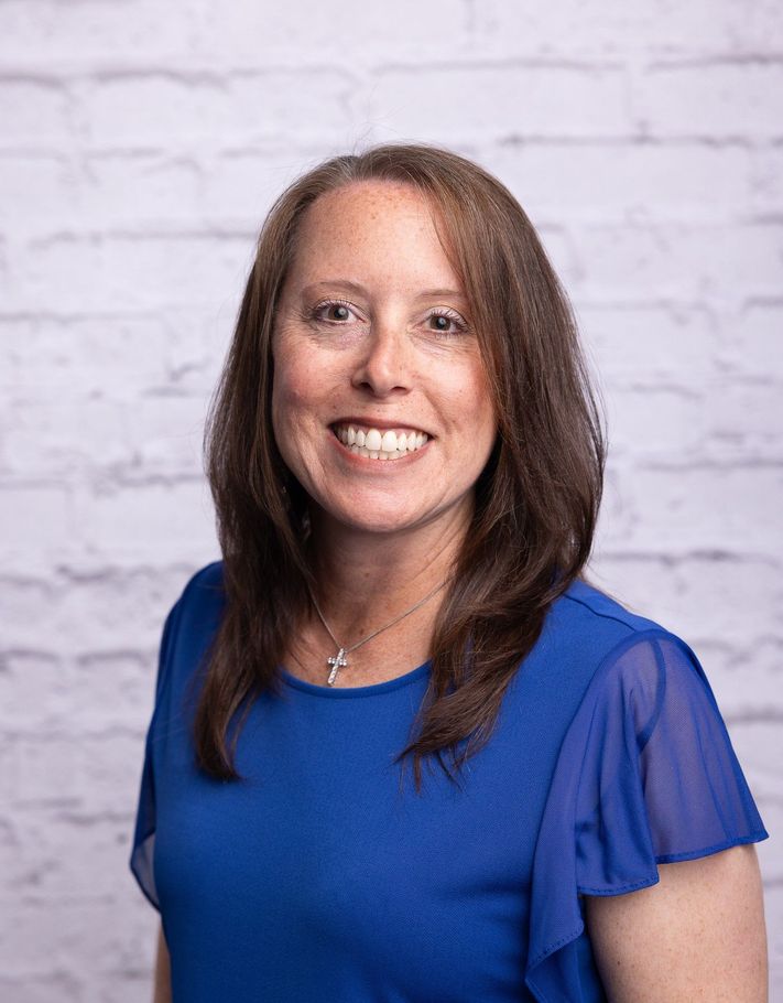 A woman in a blue shirt is smiling in front of a white brick wall.