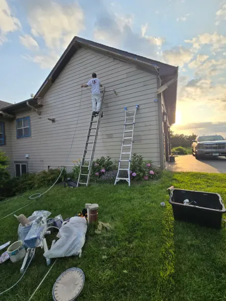 Person on ladders painting a beige house exterior. Green grass, blue sky, tools scattered below.