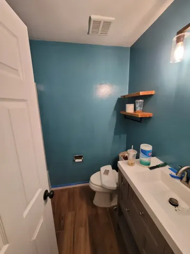 Blue-walled bathroom with wooden floor, white door, vanity, toilet, and floating shelves.