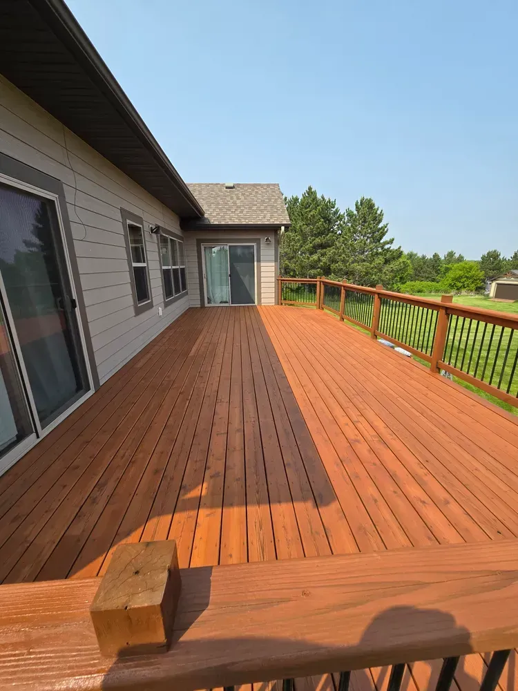 Spacious, stained wooden deck extending from a tan house; red railing, blue sky, trees in the distance.