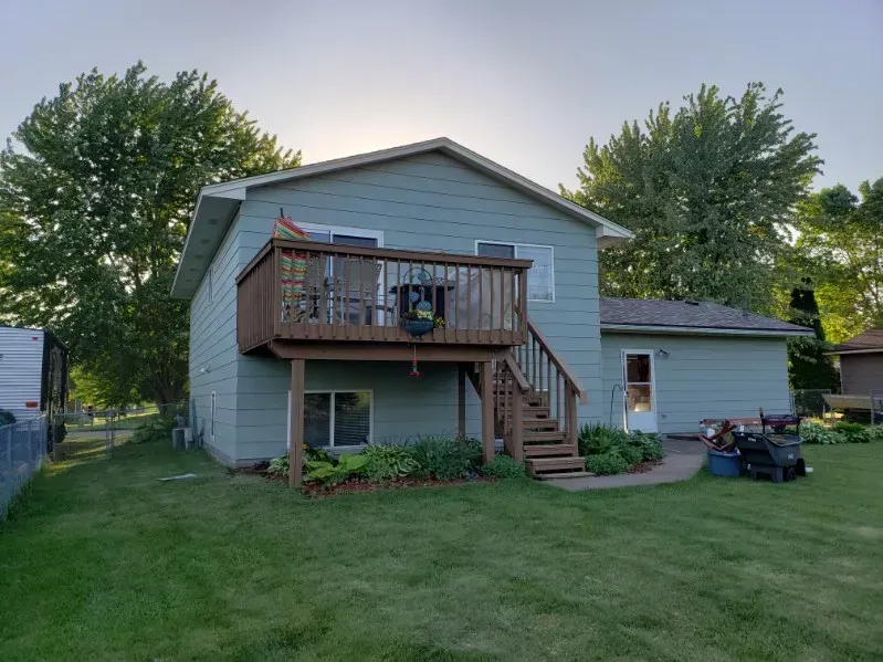 Two-story house with a brown deck. Green siding, brown stairs, and a yard with green grass.