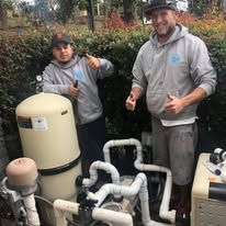 Two men are standing next to a pool pump and giving a thumbs up.
