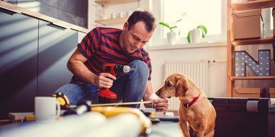 A man is kneeling down next to a dog while using a drill in a kitchen.