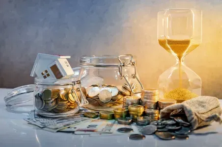 A glass jar filled with coins and a small house next to an hourglass.
