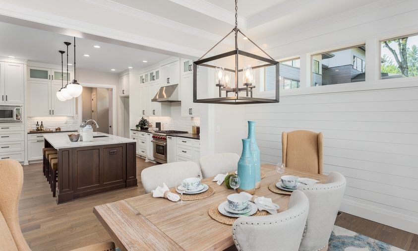 A dining room with a wooden table and chairs and a kitchen in the background.
