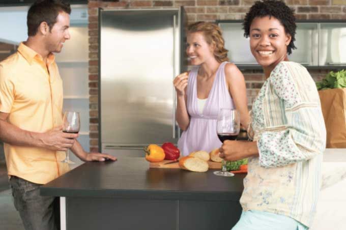 A group of people are standing around a kitchen counter drinking wine and eating vegetables.