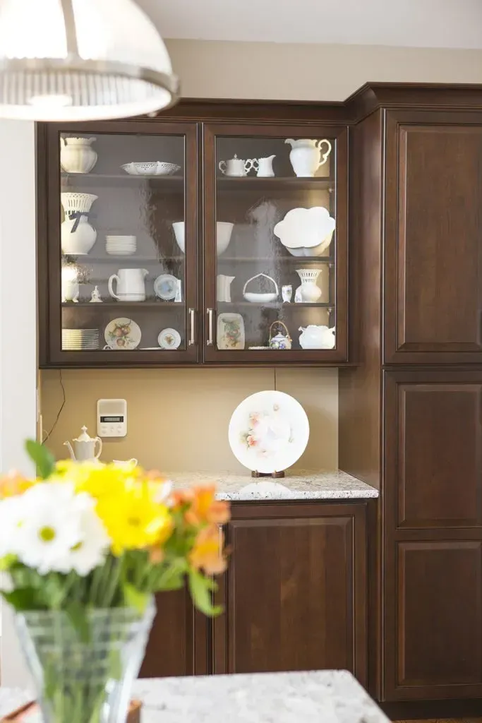 A kitchen with a glass cabinet and a vase of flowers in the foreground.