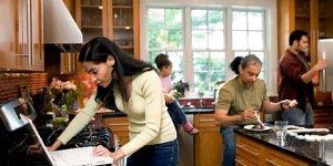 A woman is using a laptop in a kitchen while a man and a child sit in the background.