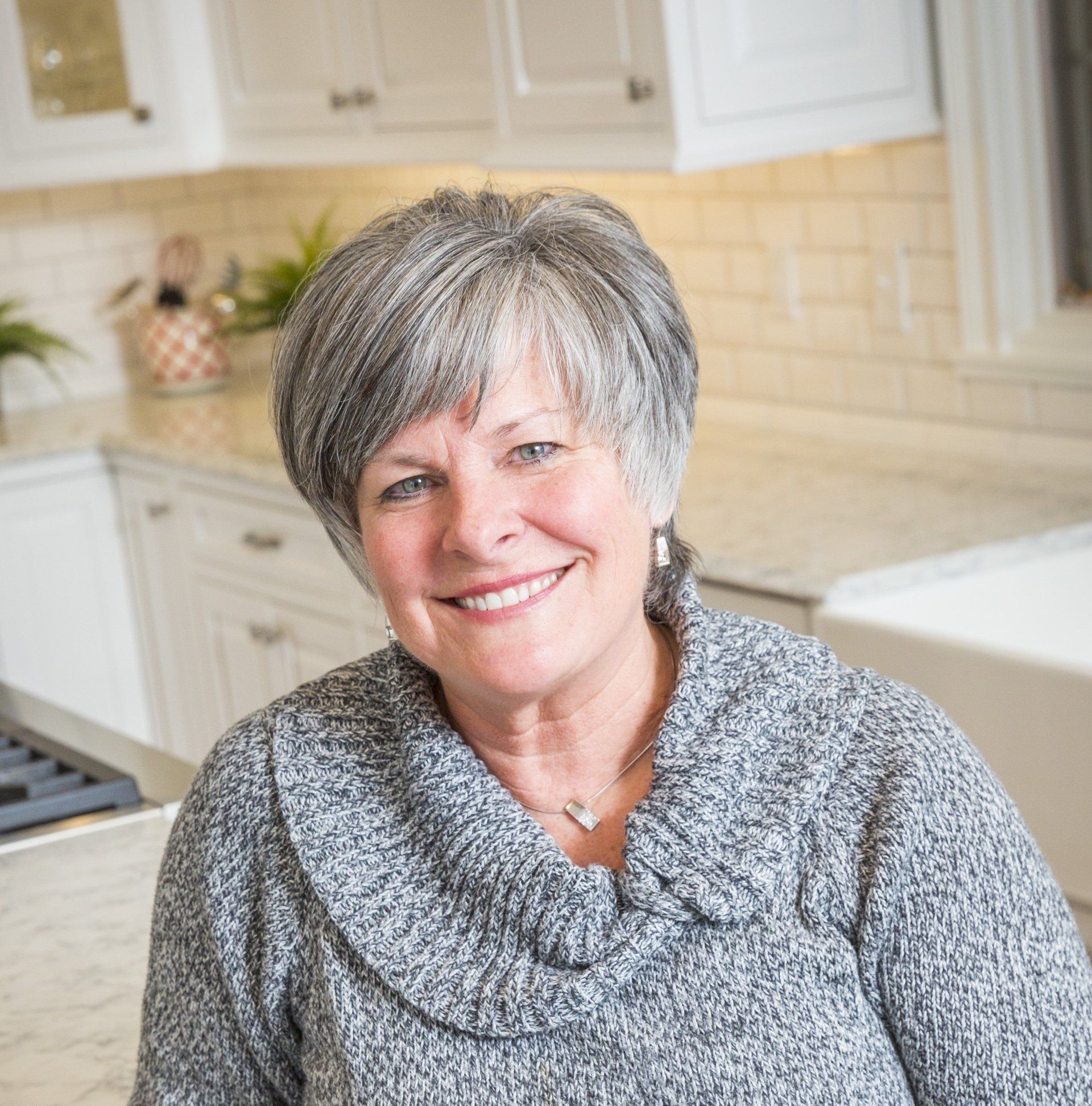 A woman in a grey sweater is smiling in a kitchen.