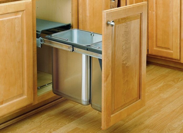 A stainless steel trash can is pulled out of a wooden cabinet in a kitchen.
