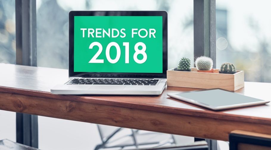 A laptop computer is sitting on a wooden desk with the words trends for 2018 on the screen.