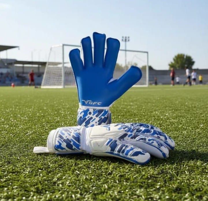 Guantes de portero azules y blancos en un campo de fútbol de césped, con jugadores y una portería al fondo.