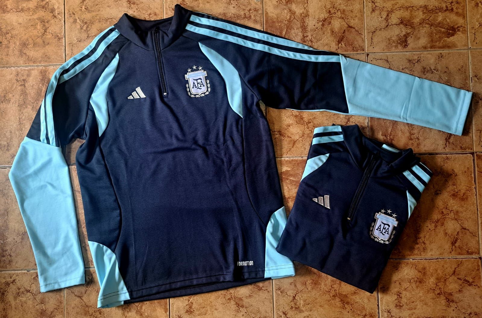 Dos sudaderas deportivas Adidas de color azul oscuro con detalles en azul claro y el logotipo del equipo, extendidas sobre un suelo de baldosas.