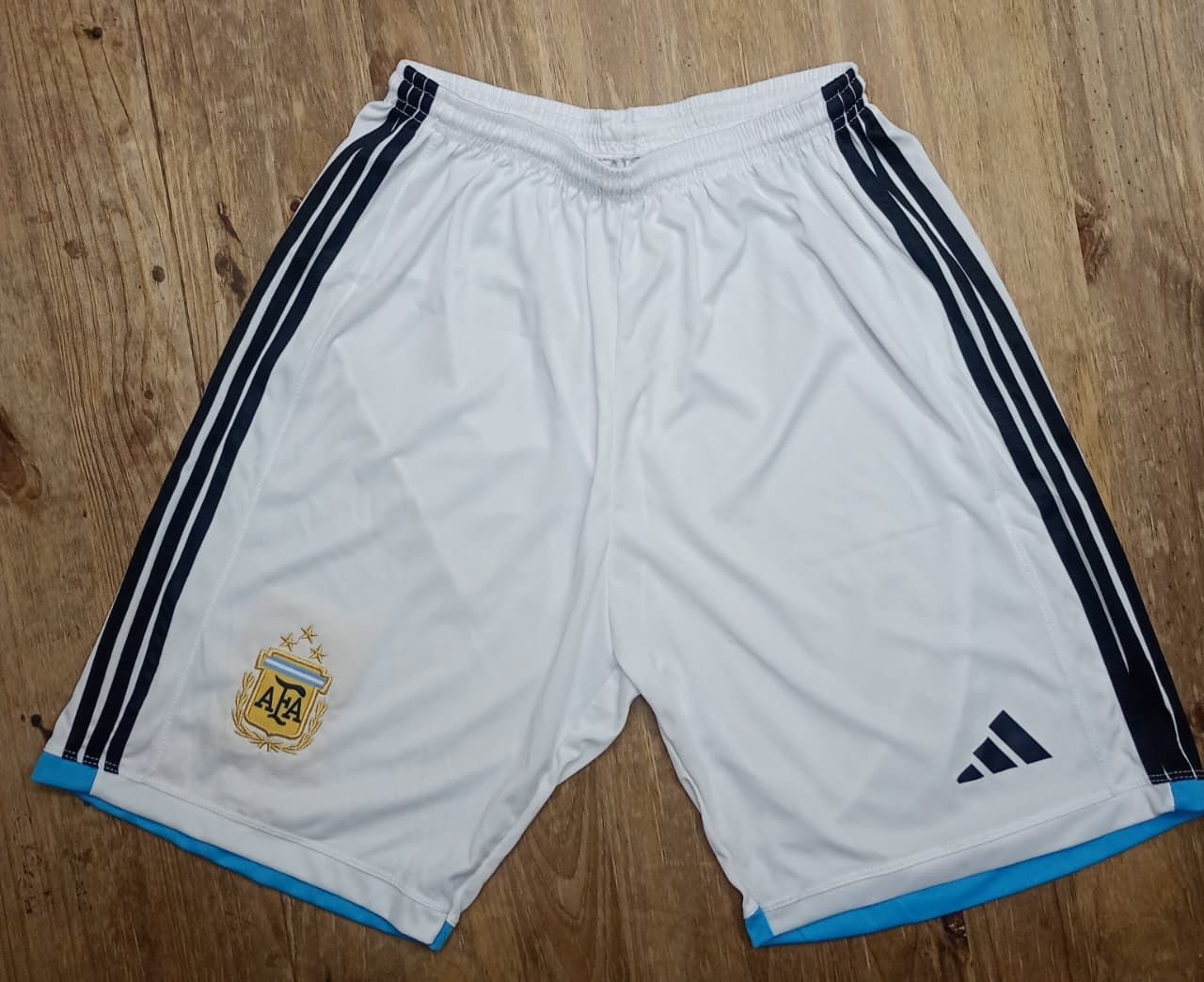 Pantalones cortos blancos de la selección argentina de fútbol con rayas azul marino, el logotipo del equipo y el logotipo de Adidas.
