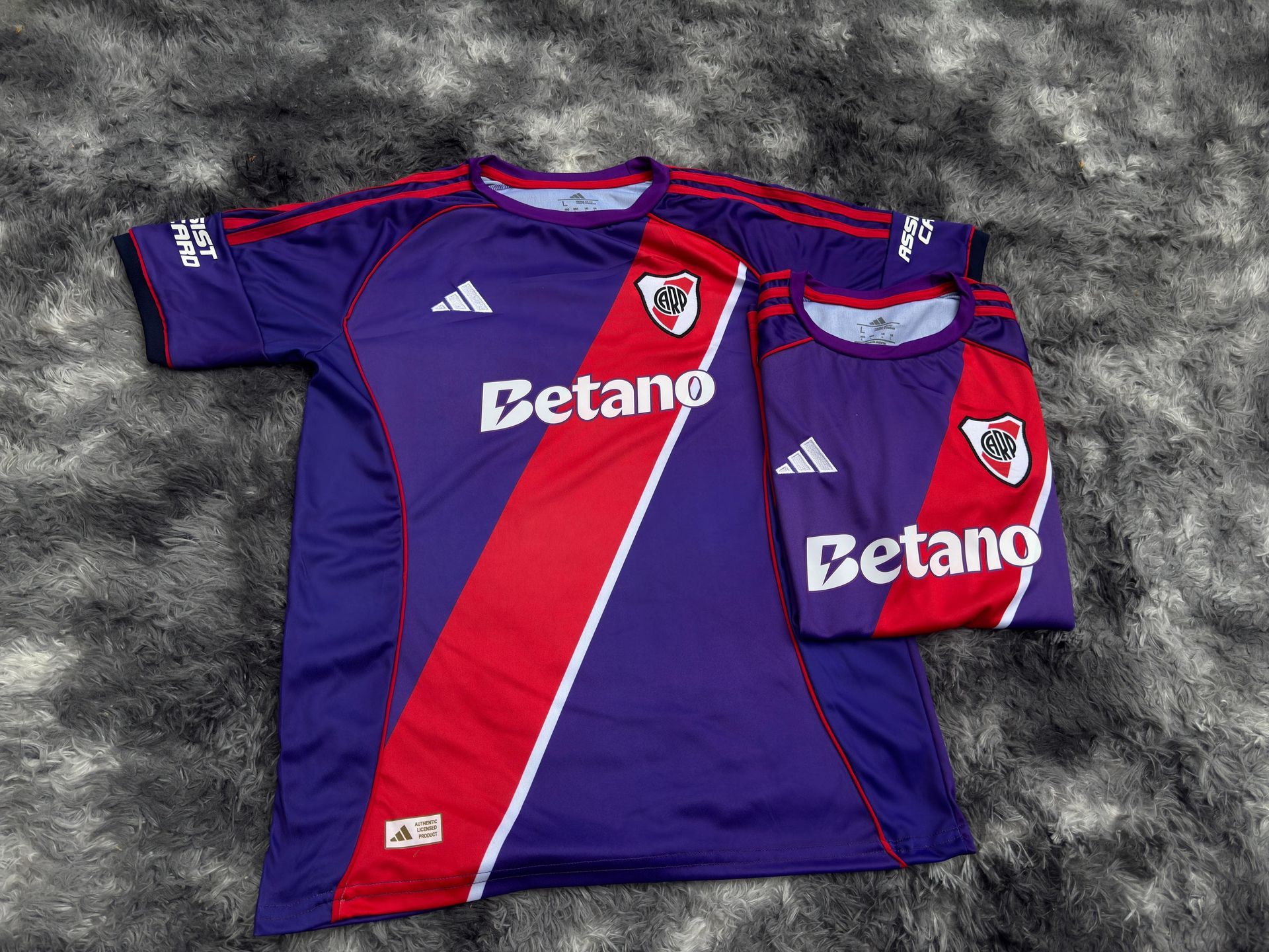 Dos camisetas moradas de River Plate con rayas diagonales rojas y logotipos blancos de Betano sobre una alfombra gris de pelo largo.