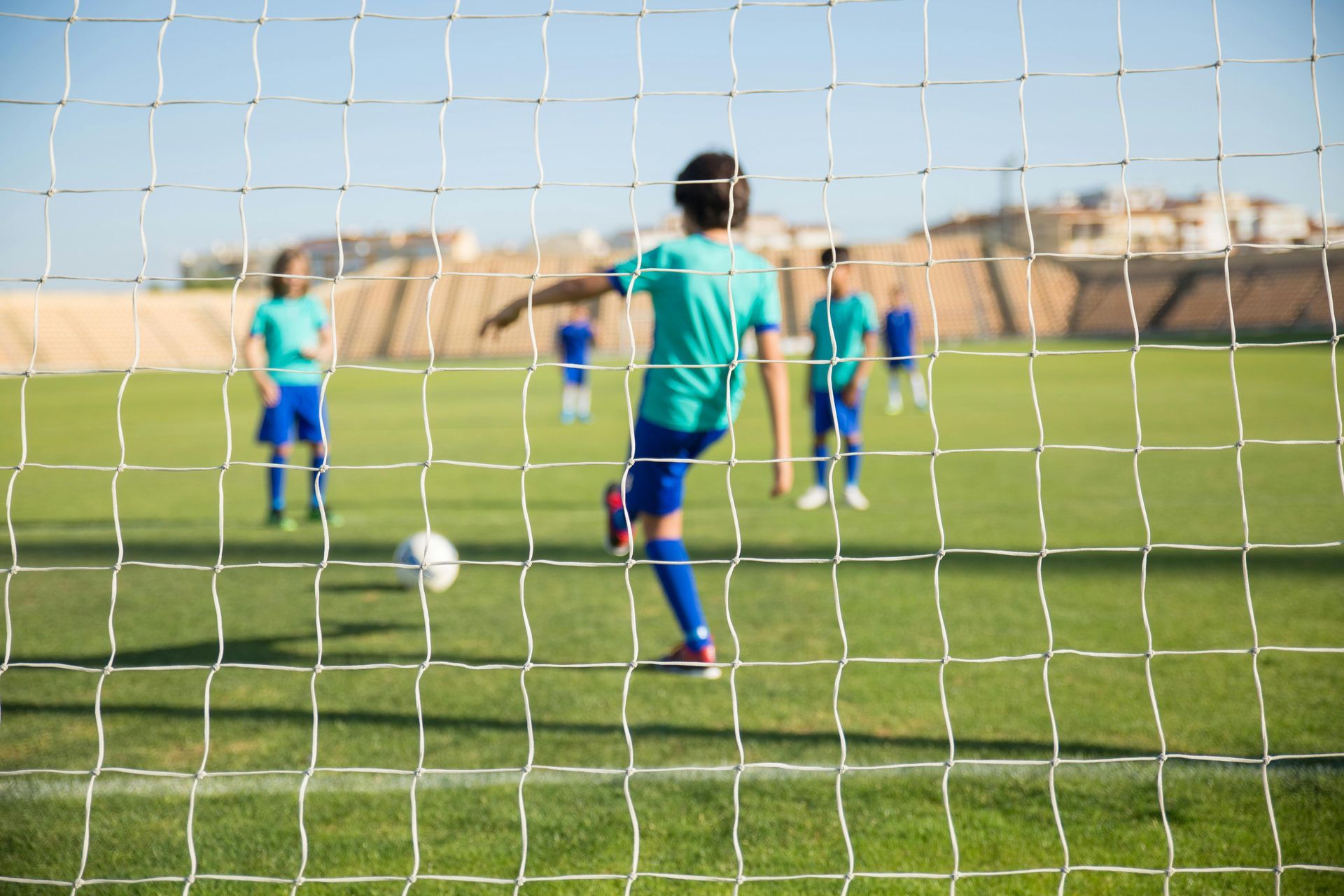 Jugador de fútbol patea el balón hacia la red, otros jugadores en el fondo en el campo verde.