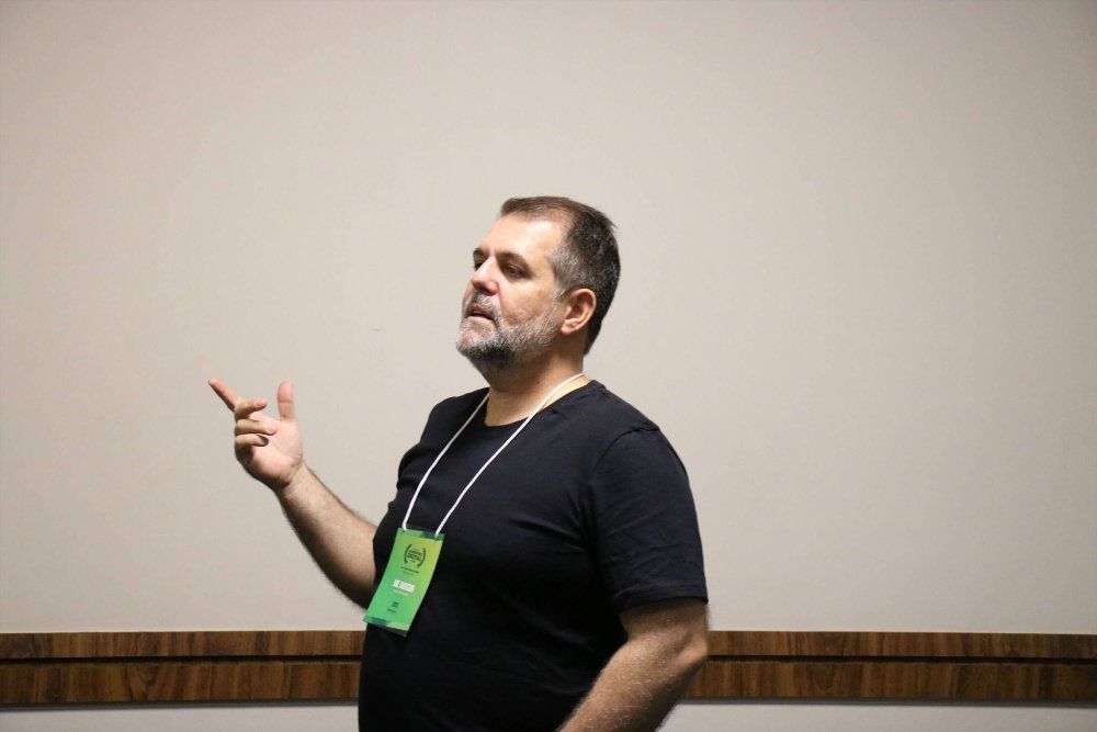 Um homem com barba está parado em frente a uma parede branca e apontando.