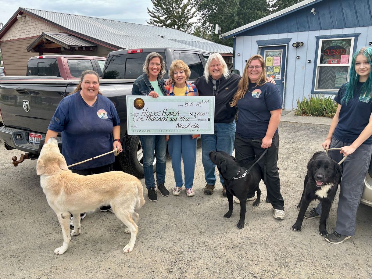 Group of people holding a large check for an animal shelter, with three dogs. The setting appears to be outdoors at a small building.