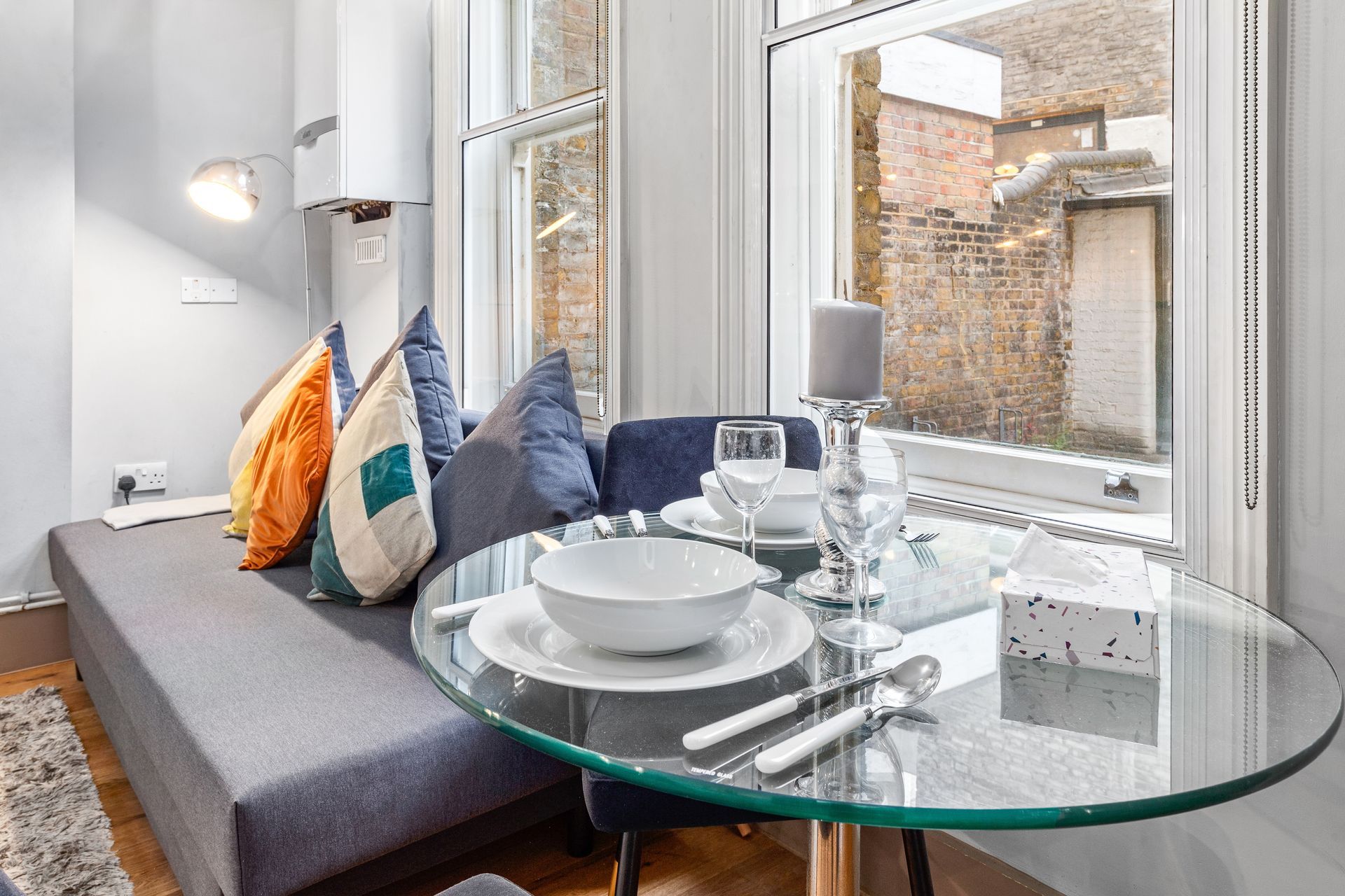 Cozy dining nook with a glass table set for a meal, next to a window and a cushioned bench with pillows.
