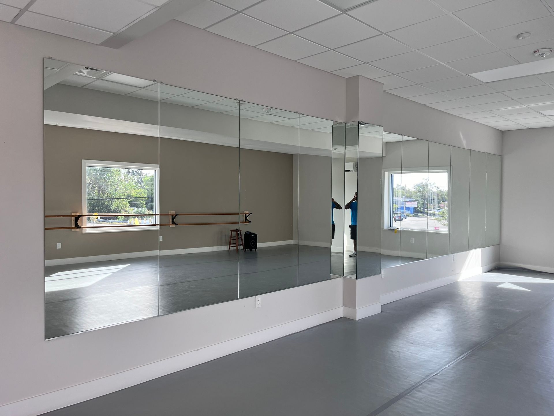 Dance studio interior with wall-mounted mirrors, barre, gray floor, and natural light from windows.