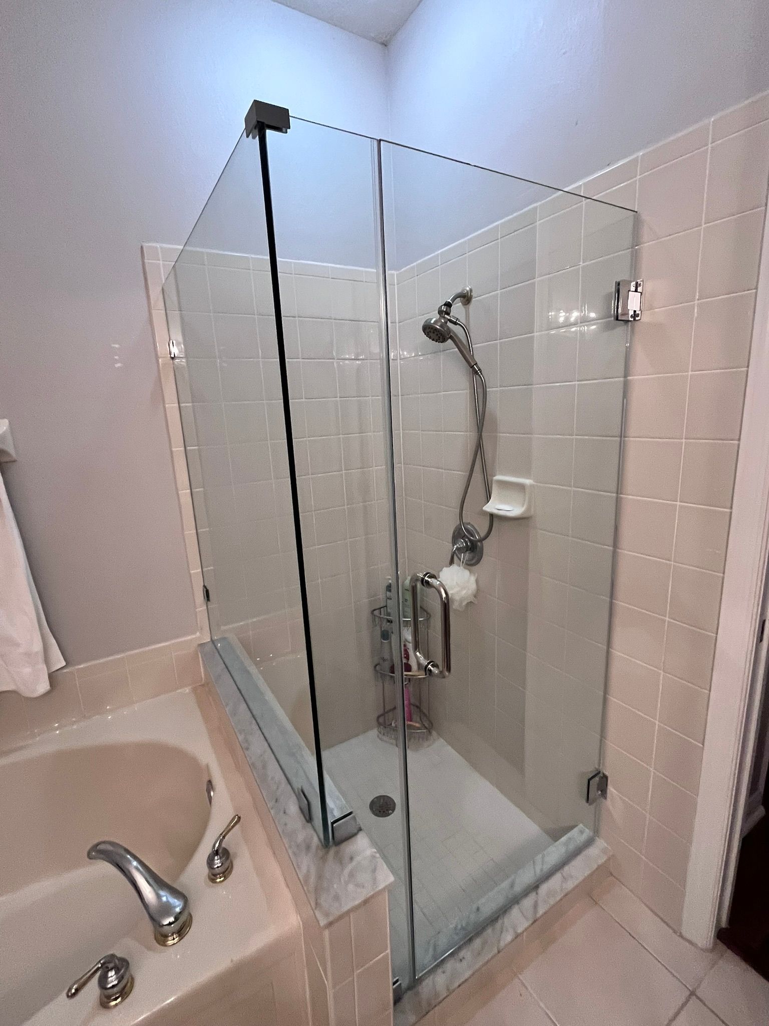 Shower stall with clear glass doors and a shower head, next to a bathtub. Beige tile.