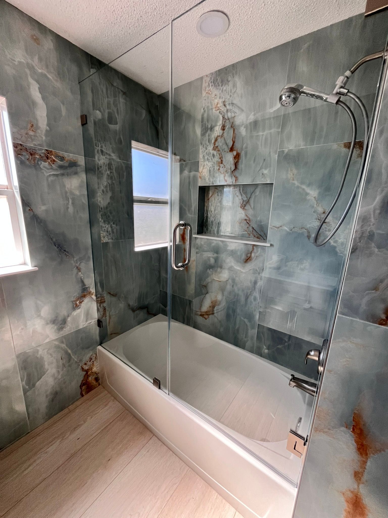 Bathroom with blue-green marble tiles, glass shower door, white bathtub, and silver shower fixtures.