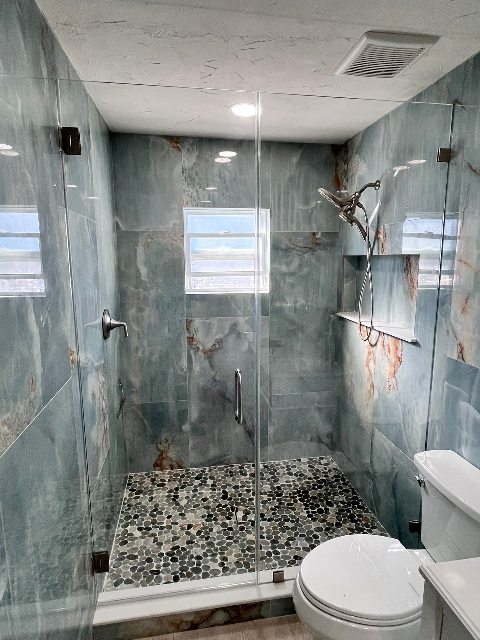 Modern shower with blue-tiled walls, glass door, and pebble floor. Includes toilet and small window.