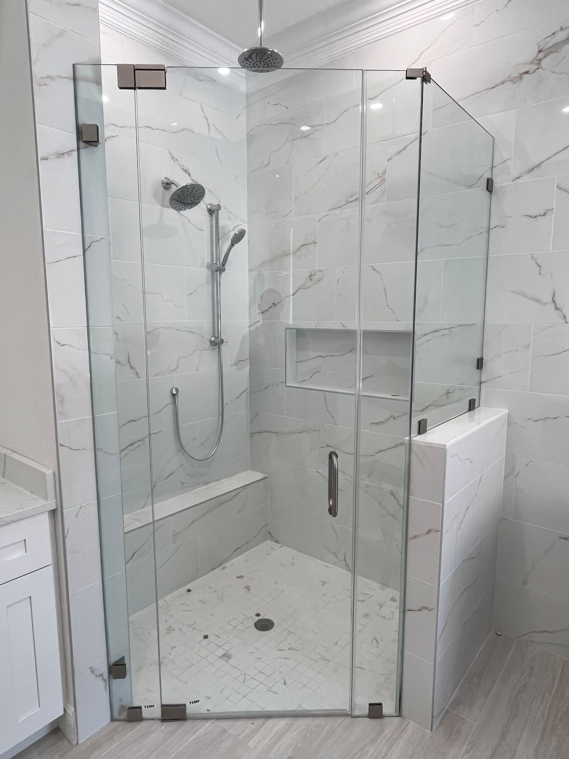 Glass-enclosed shower with white marble-like tile. Includes a bench, shower head, and recessed shelf.