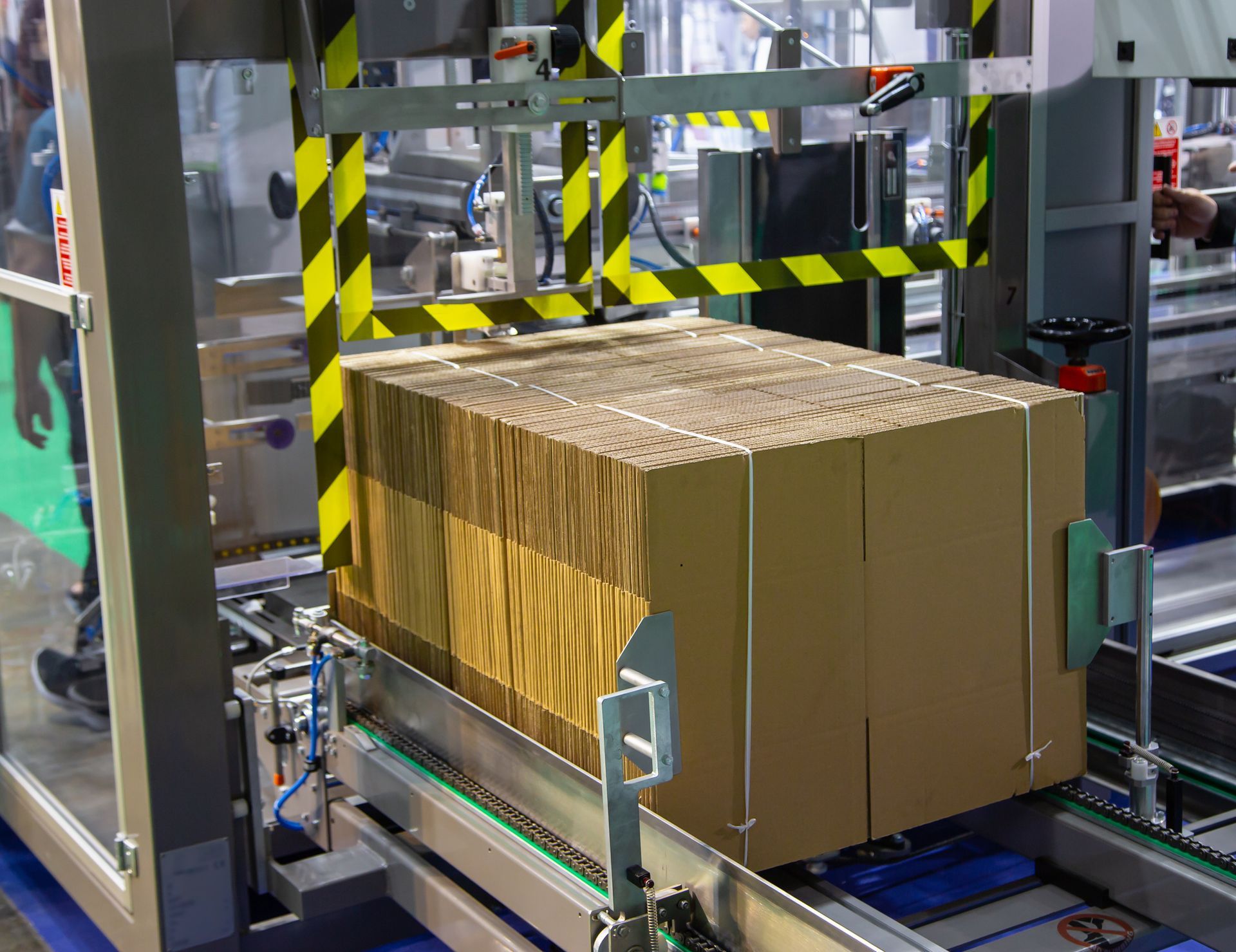 Boxes stacked on a conveyor belt in a factory setting. Yellow and black safety bars.