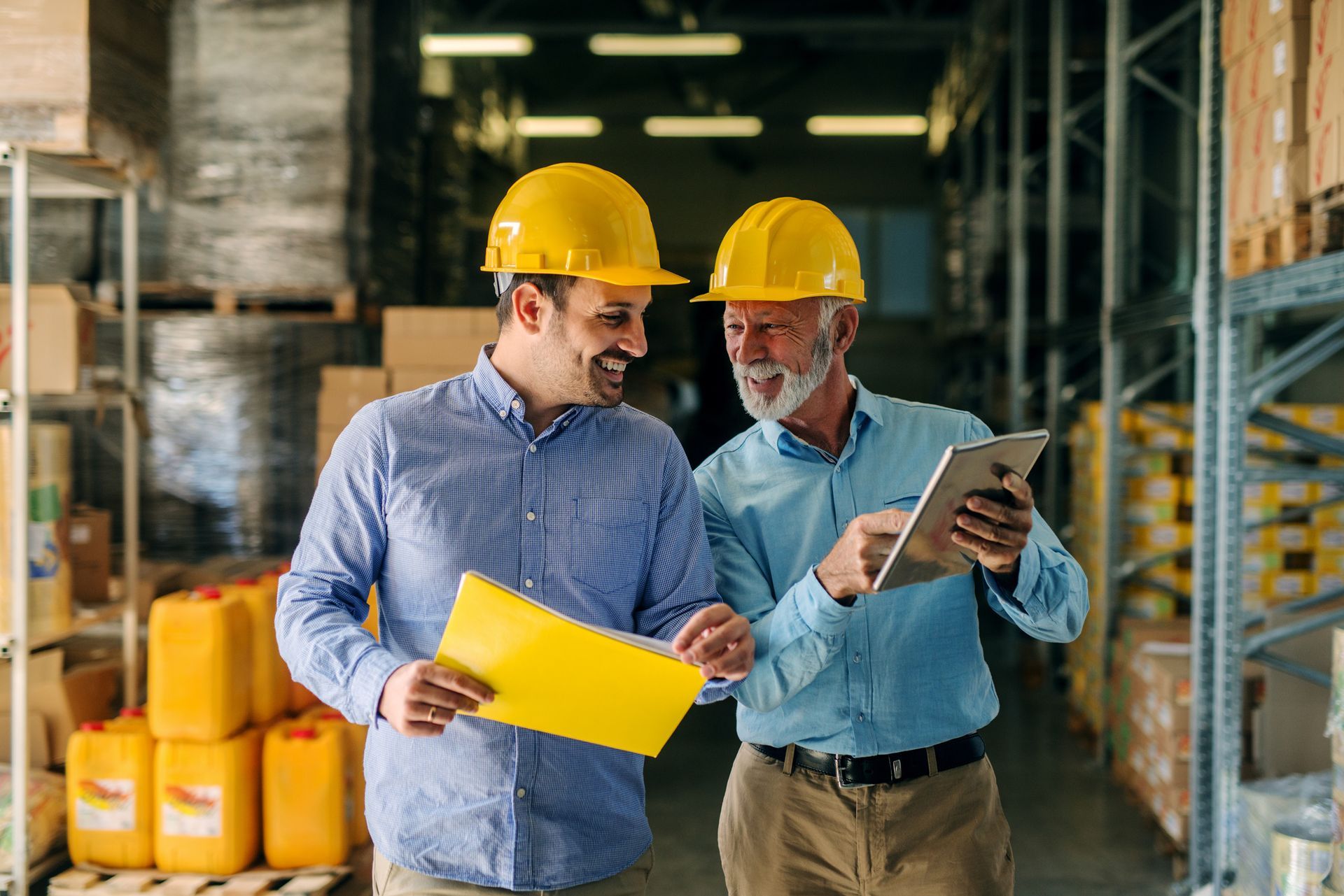 Two men in hard hats and button-down shirts discuss paperwork in a warehouse.
