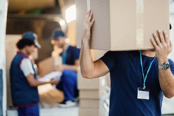 Delivery worker carrying a large box on their shoulder; two other workers in background at a delivery van.