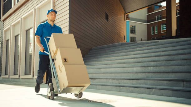 Delivery person in blue uniform rolls a dolly with boxes up stairs near a building.