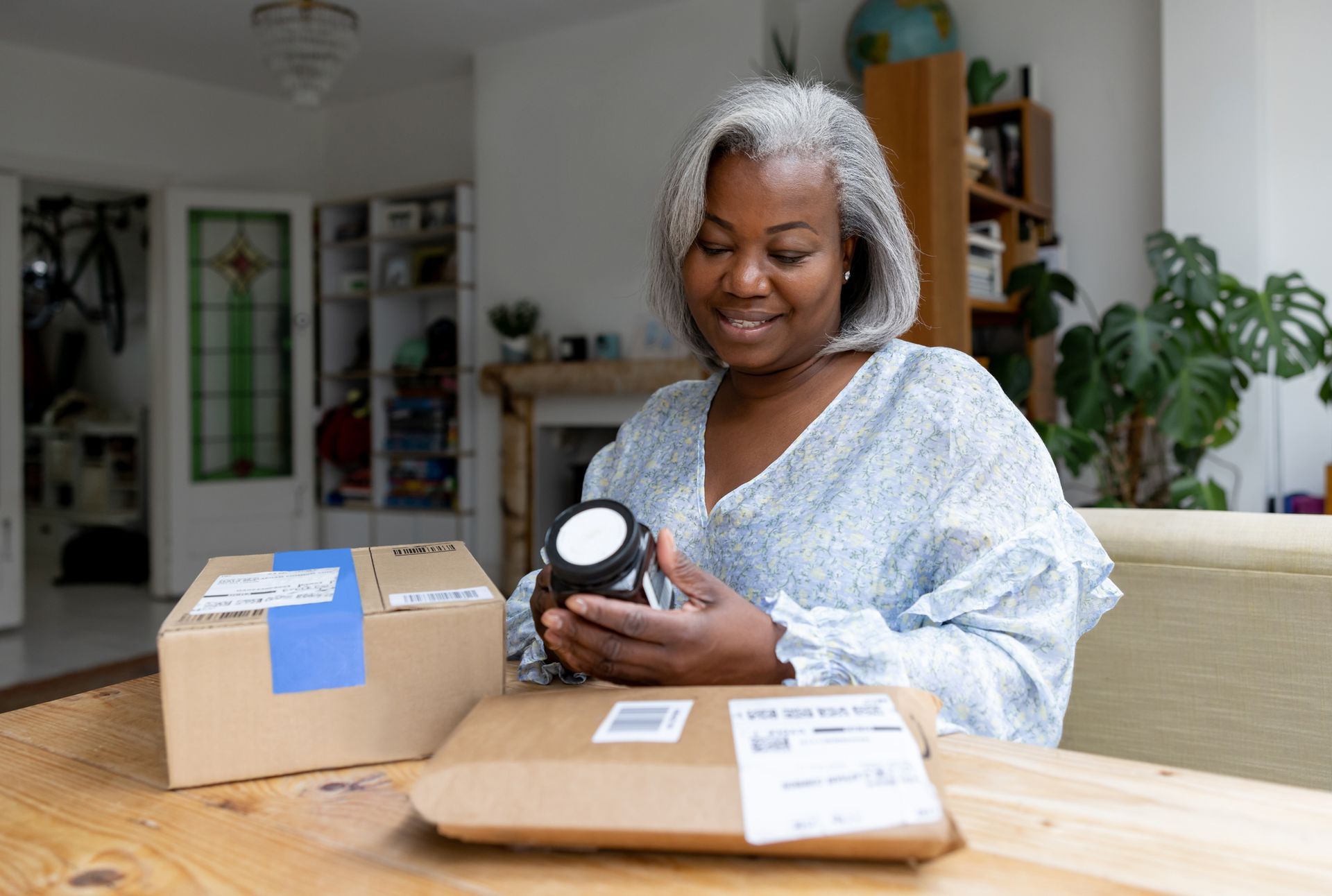 Woman with gray hair smiles, holding an object, parcels on table. Interior setting with natural light.