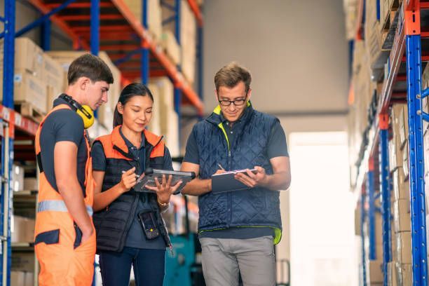 Three warehouse workers reviewing inventory; woman holding tablet, man writing, another man wearing high visibility gear.