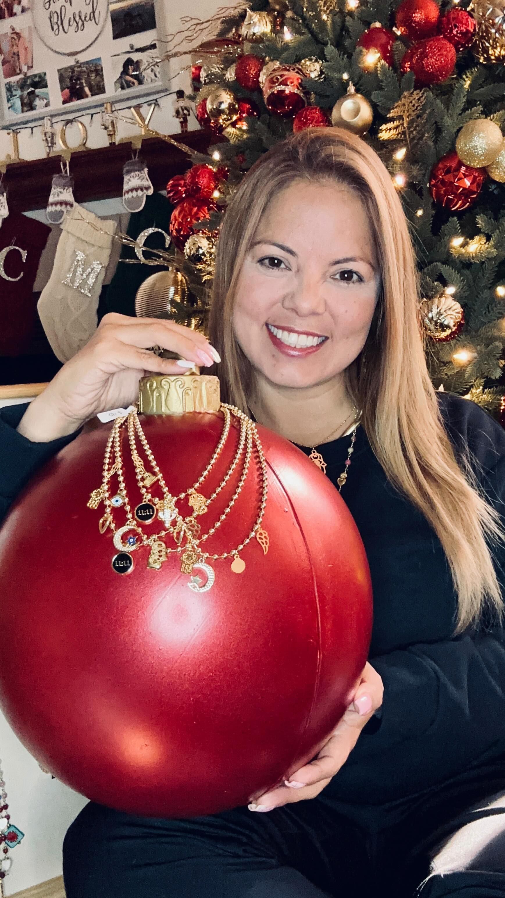 A woman is holding a large red christmas ornament in front of a christmas tree.