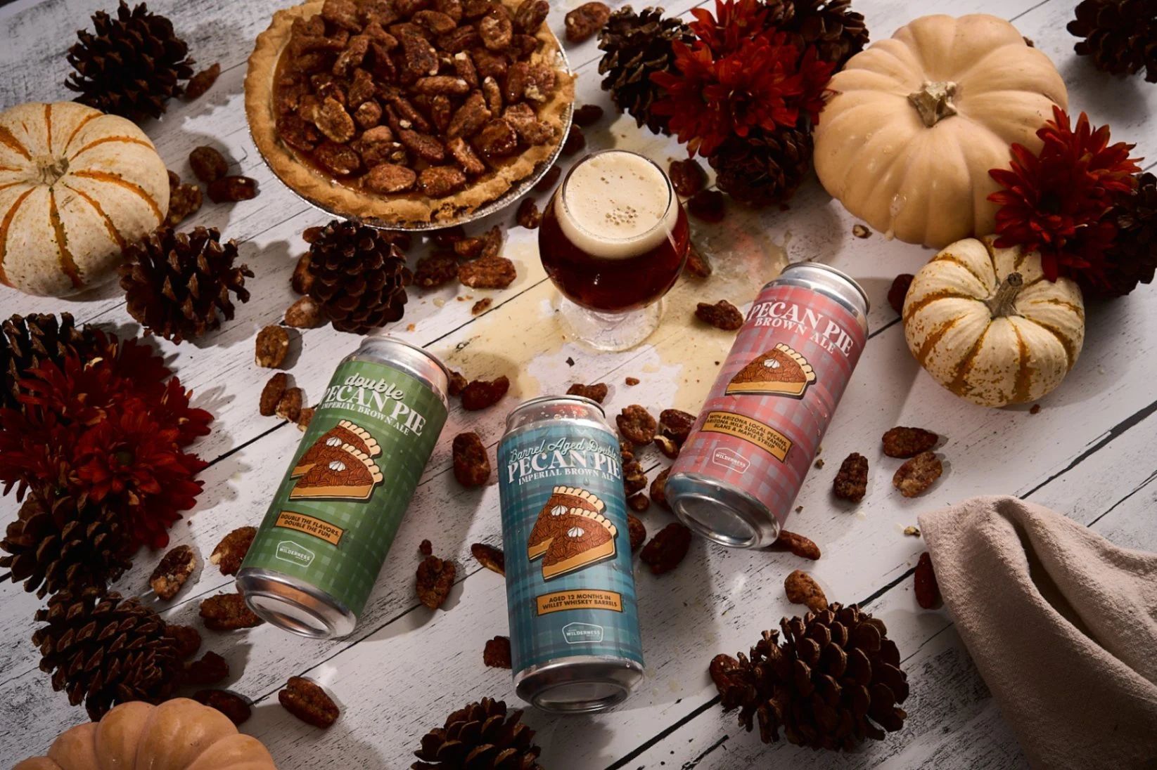 A table topped with cans of beer , pumpkins , pine cones and a pie.