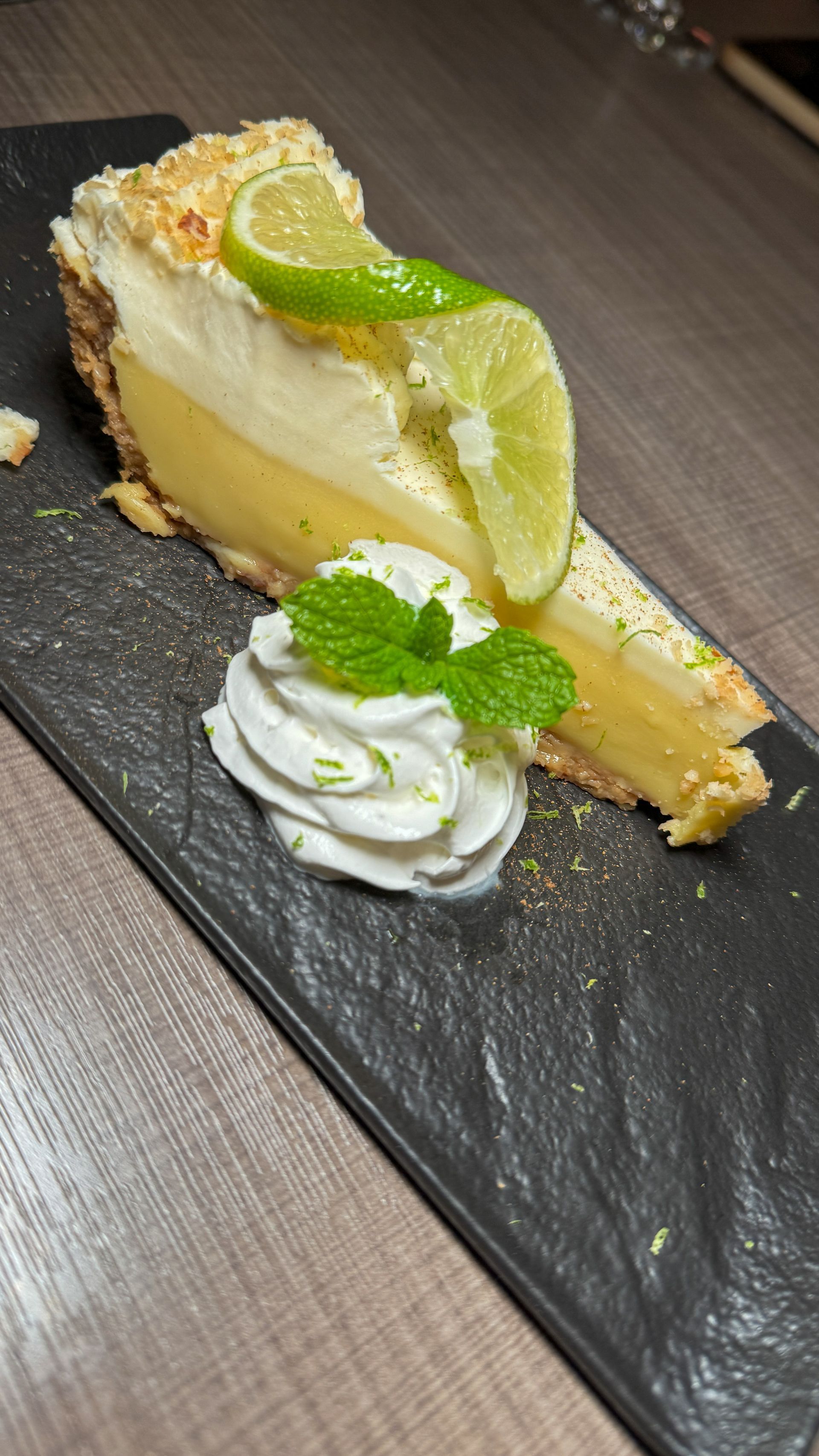A slice of key lime pie with whipped cream and mint leaves on a black plate.
