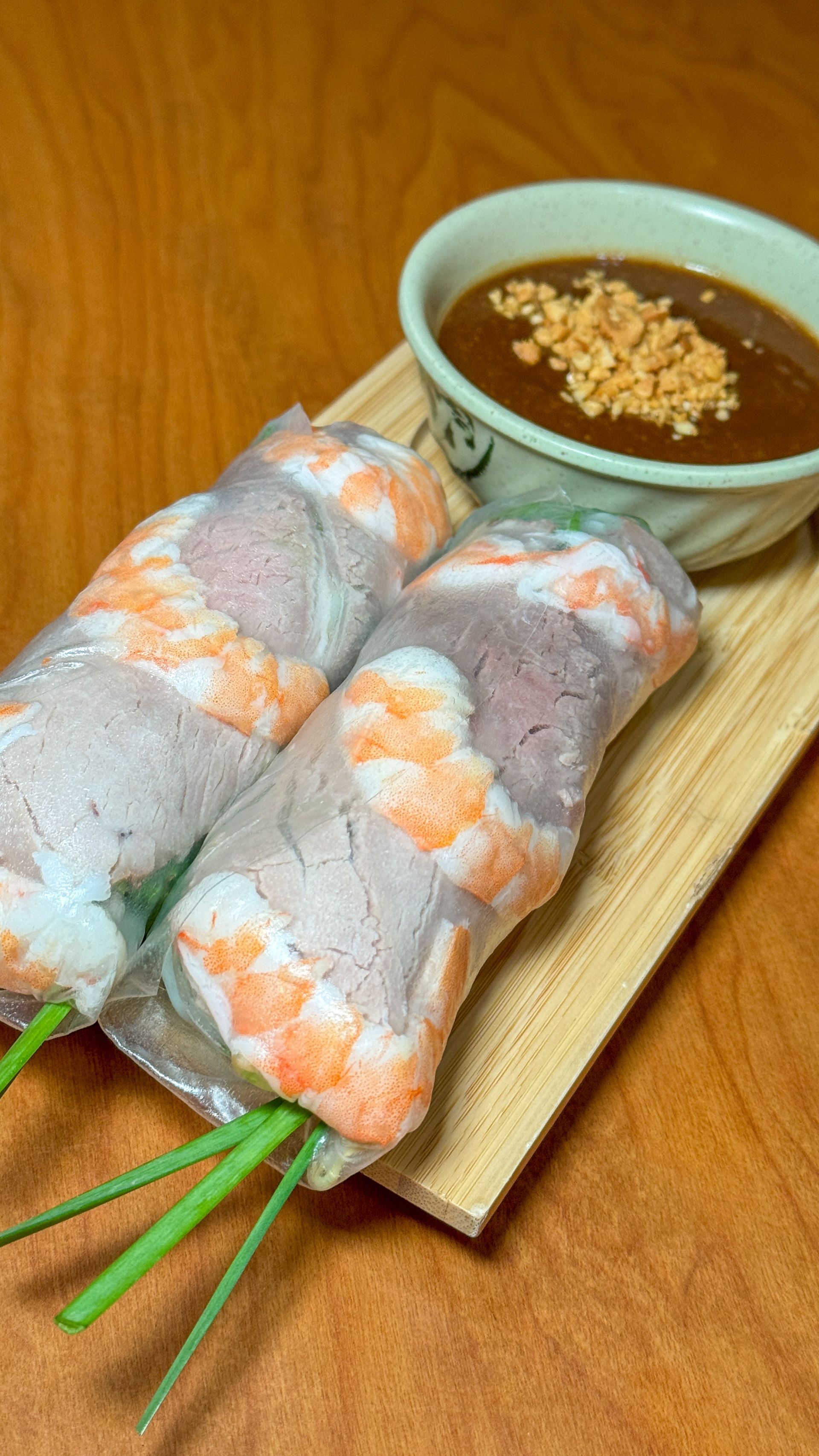 Two shrimp spring rolls are sitting on a wooden cutting board next to a bowl of dipping sauce.
