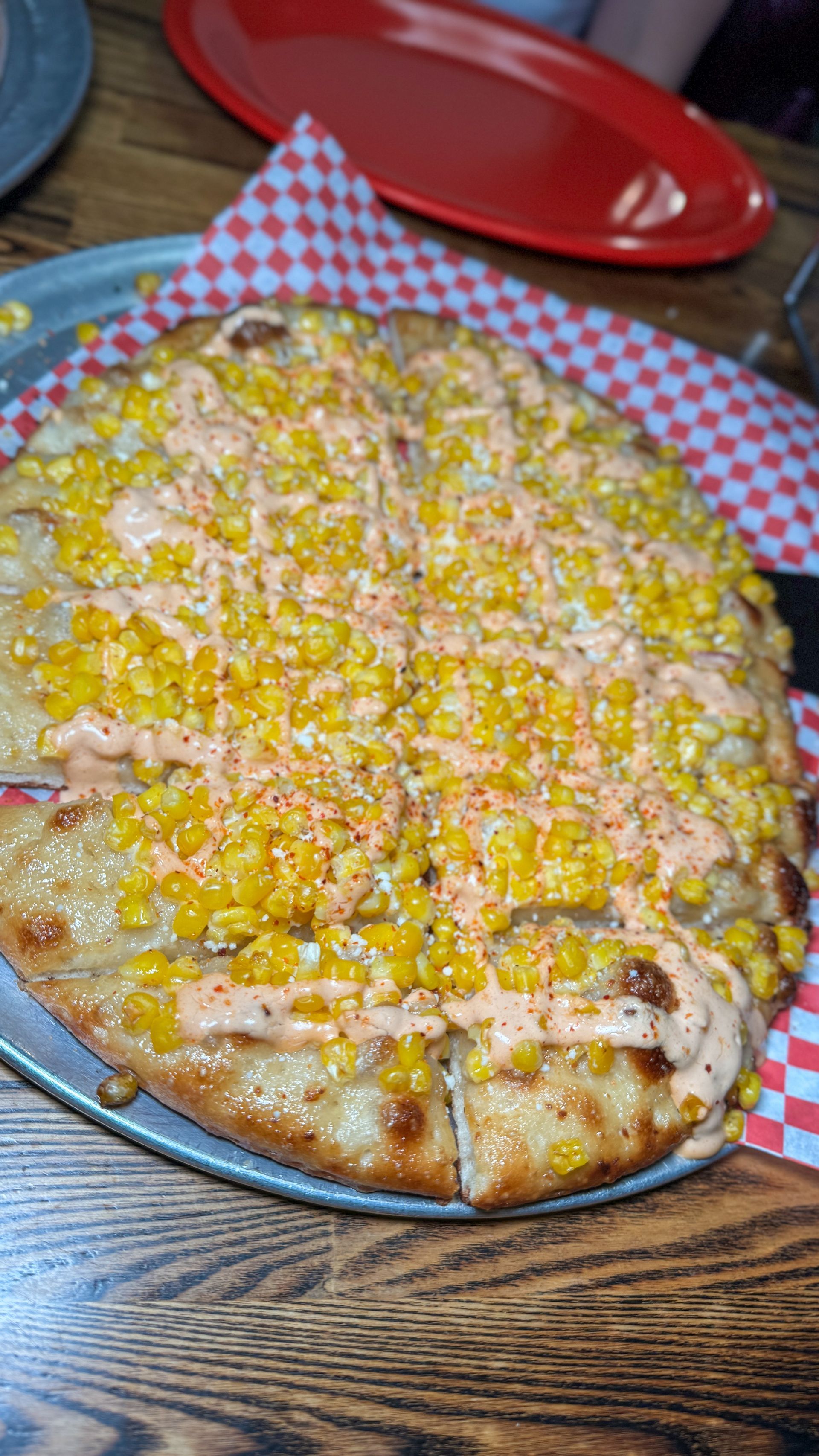 A pizza with corn on top is on a pizza pan on a wooden table.