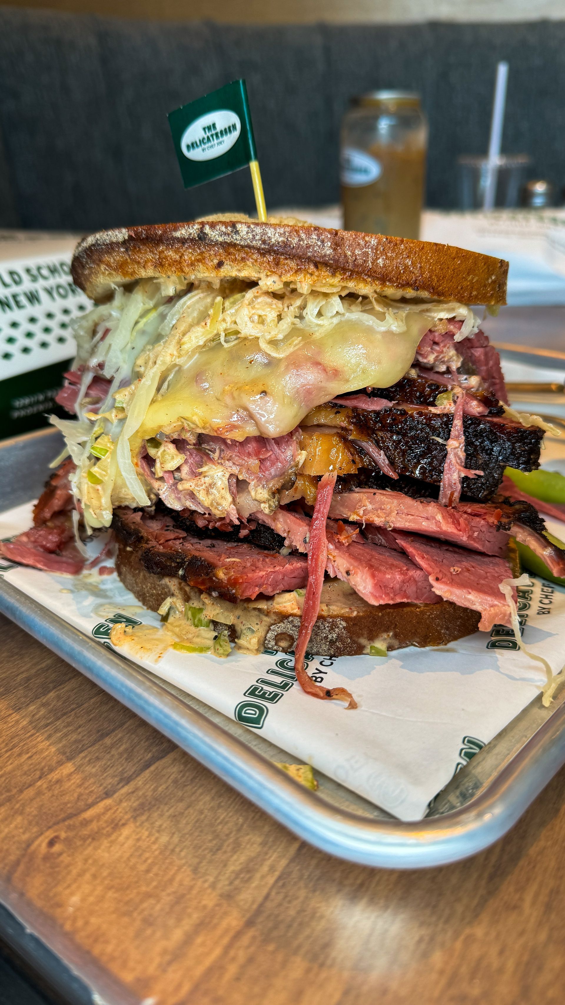 Pastrami sandwich on rye with cheese, sauerkraut, and a toothpick flag on a metal tray.