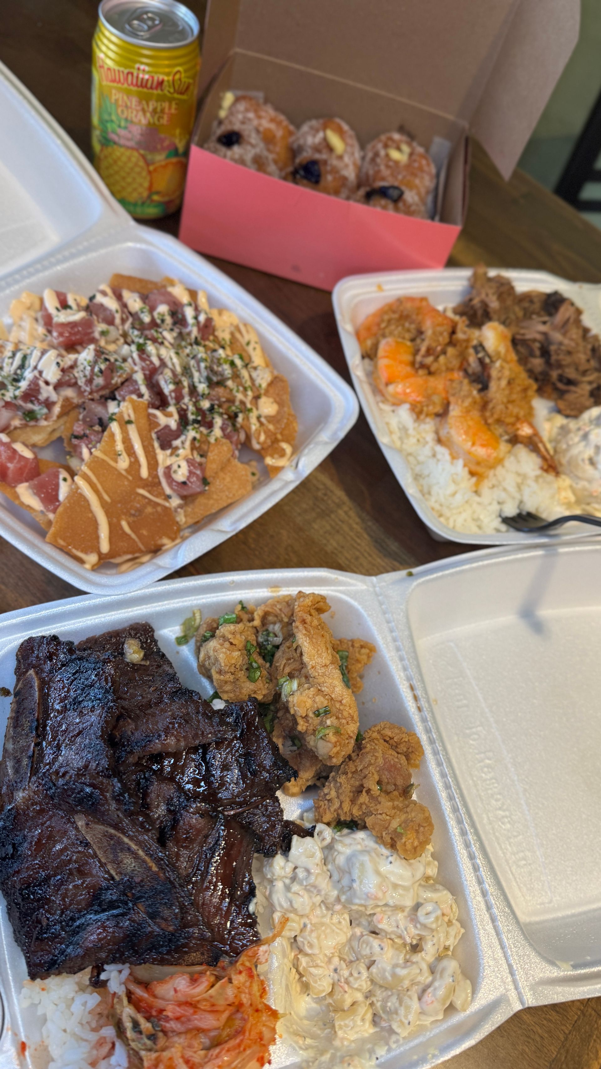 Takeout food, including ribs, shrimp, and nachos, in styrofoam containers with a drink and donuts.