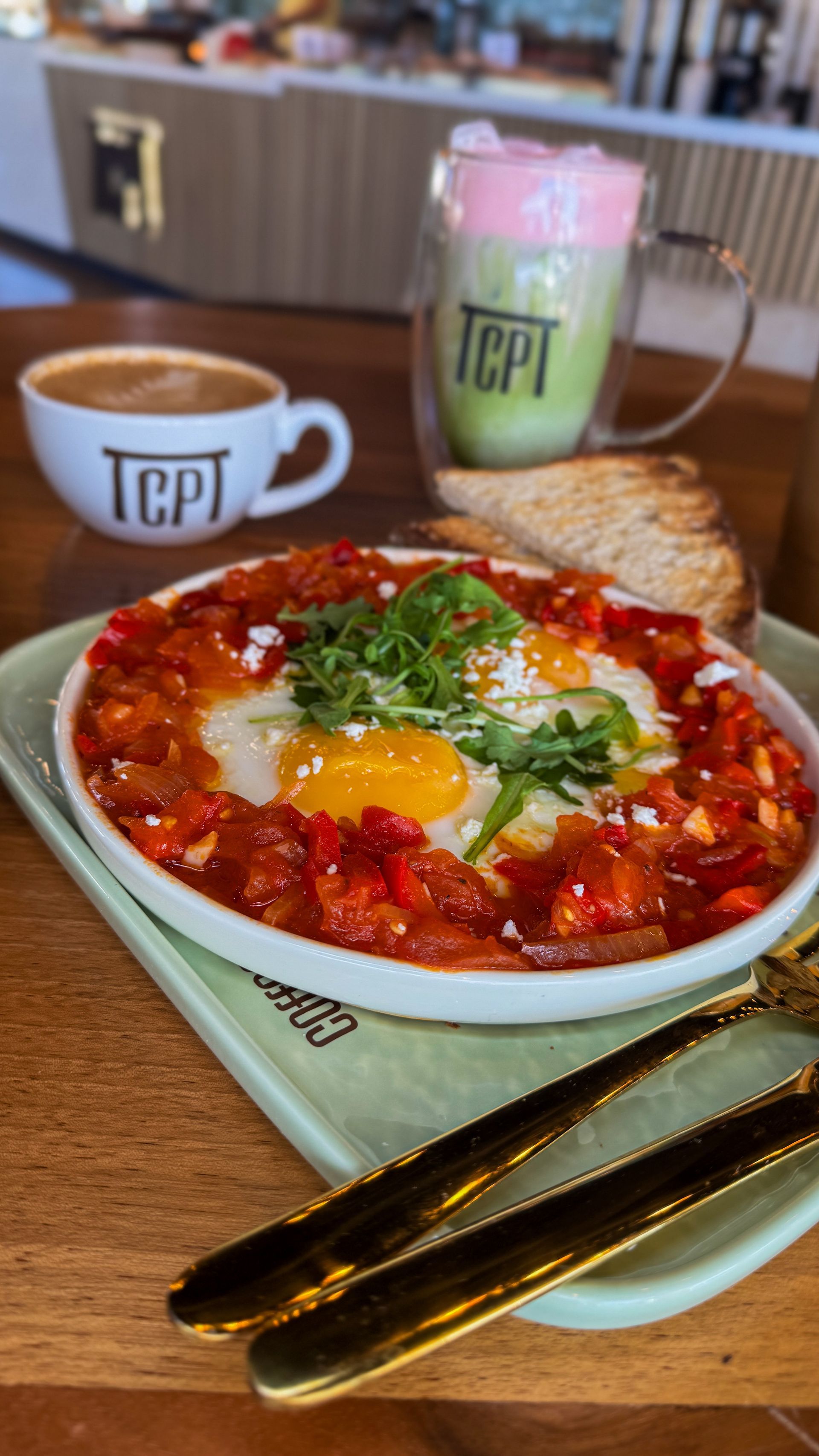 Breakfast plate with eggs in tomato sauce, coffee, matcha latte, and toast.
