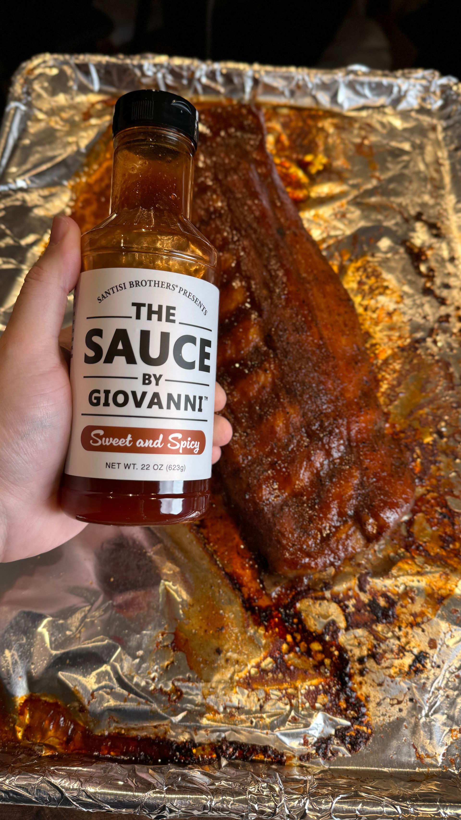 A person is holding a bottle of sauce in front of a pan of ribs.