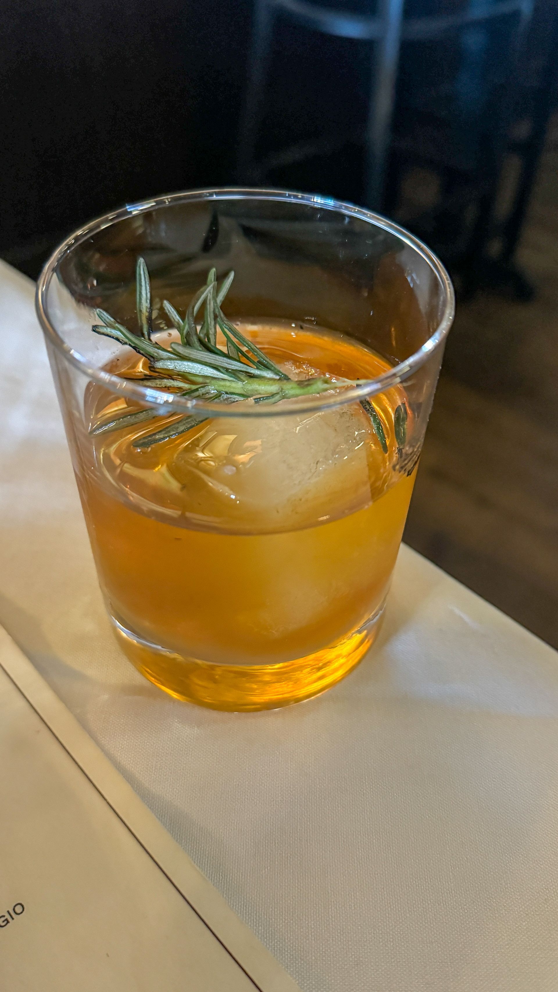 A glass of whiskey with ice and rosemary on a table.