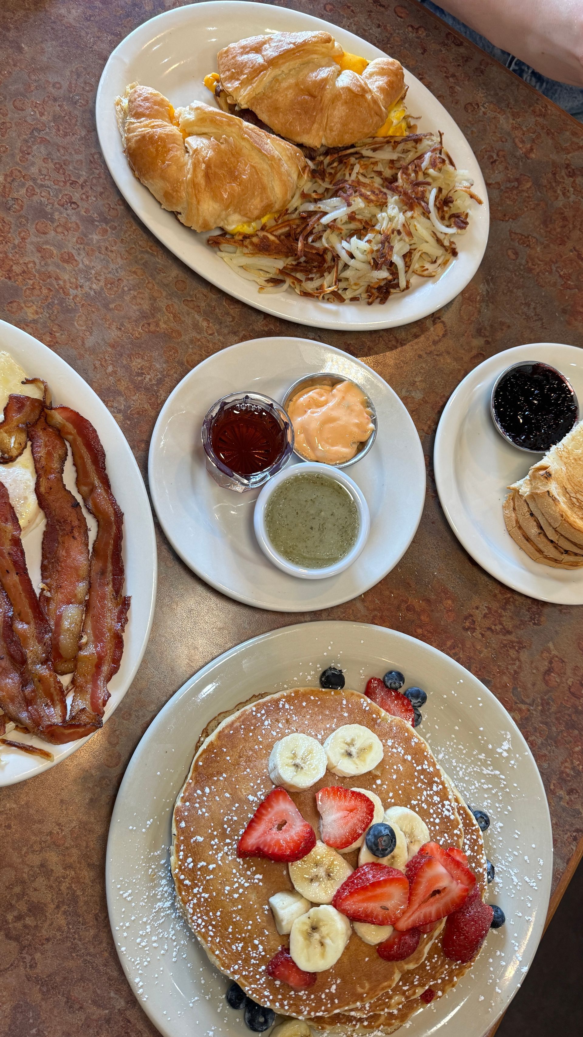 A table topped with plates of food including pancakes , croissants , bacon and eggs.