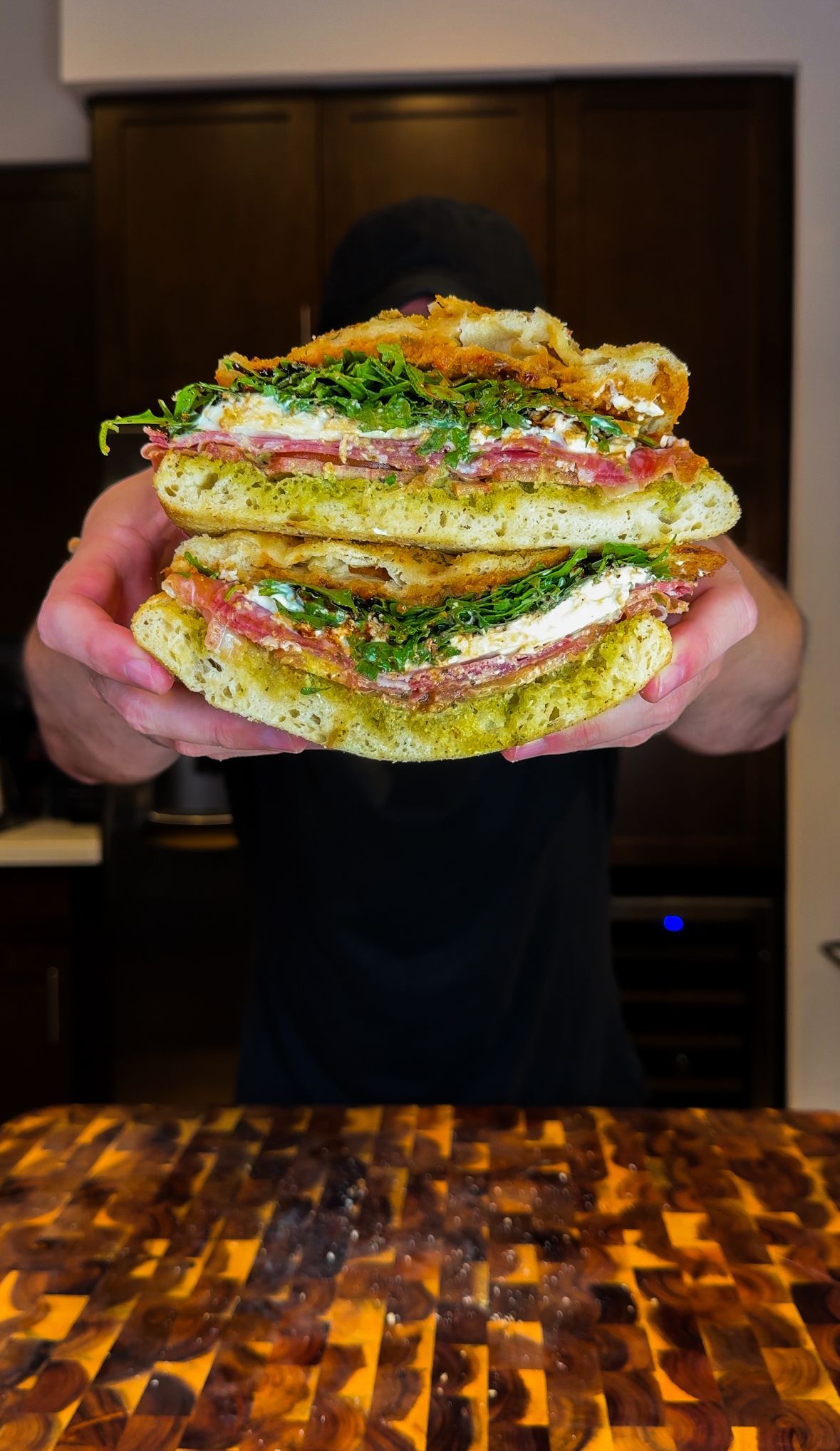 Person holding a large, layered sandwich with salami, arugula, and pesto on a wooden cutting board.