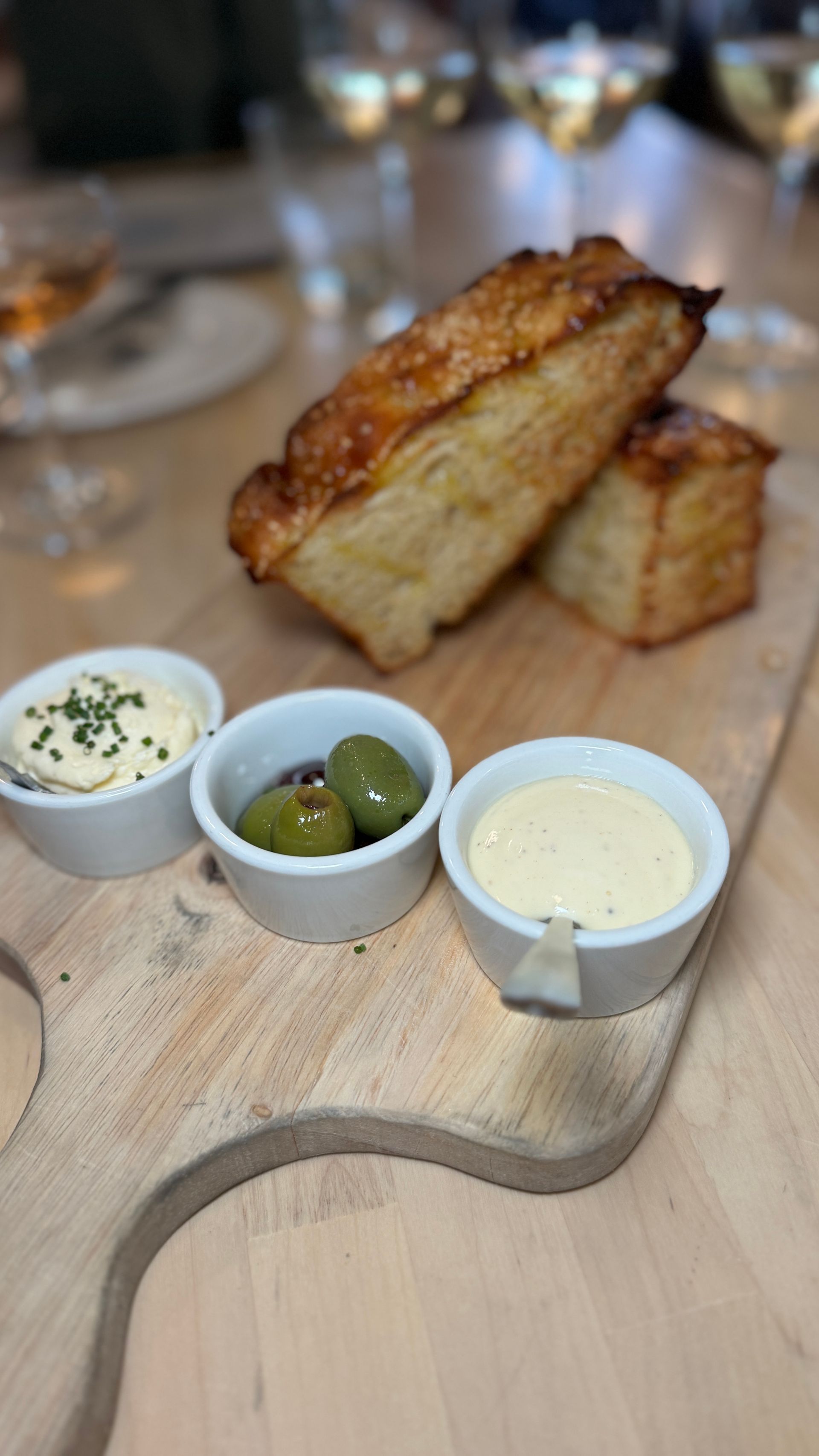 A wooden cutting board with bread , olives , and sauces on it.